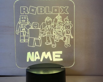 May include: A white acrylic night light with a black base. The light features a white outline of Roblox characters and the word "ROBLOX" at the top. The light also has a customizable name area that says "NAME".