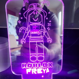 May include: A purple acrylic night light with a white outline of a Roblox character wearing a hat and holding a box. The light has the text "Roblox Freya" at the bottom.