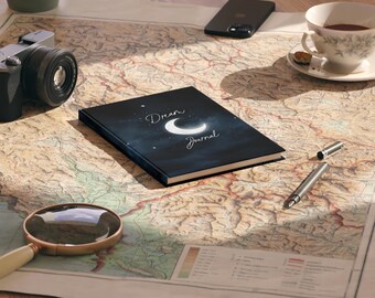 May include: A black journal with the words "Dream Journal" and a crescent moon design on the cover. The journal is on a vintage map, with a camera, magnifying glass, pen, phone, and teacup nearby.