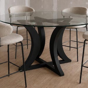 May include: A round dining table with a clear glass top and a black curved base, accompanied by four high-backed chairs featuring beige upholstered seats and thin black metal legs, arranged in a room with light wood flooring and unfinished walls.