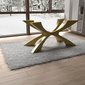 May include: A modern, gold-colored table base with a unique, curved design. The table base is set on a fluffy, light gray rug, with a snowy winter scene visible through a large window in the background. The room has light wood flooring.