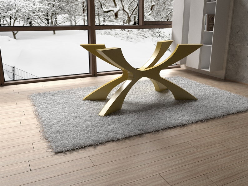 May include: A modern, gold-colored table base with a unique, curved design. The table base is set on a fluffy, light gray rug, with a snowy winter scene visible through a large window in the background. The room has light wood flooring.