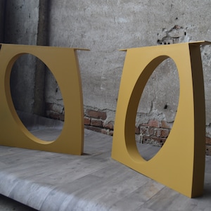 May include: Two yellow metal table legs with a rounded cutout in the center. The legs are standing on a light brown wooden surface.