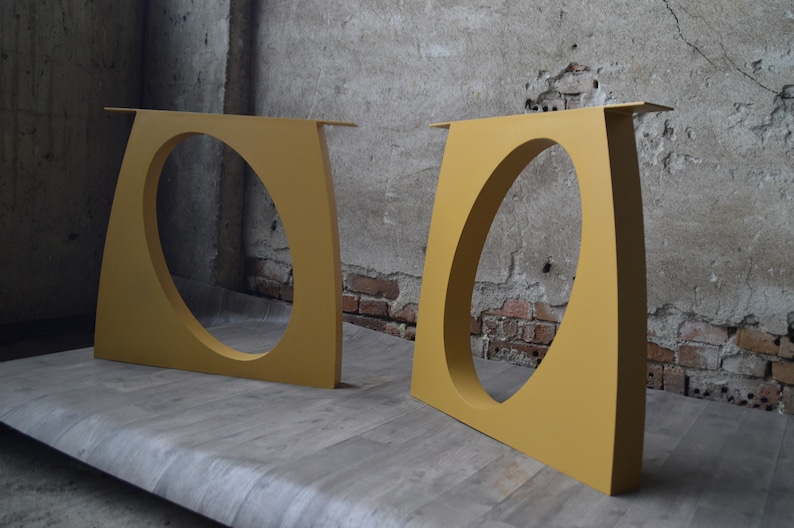 May include: Two yellow metal table legs with a rounded cutout in the center. The legs are standing on a light brown wooden surface.