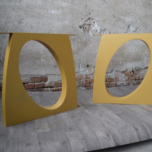 May include: Two gold metal table legs with an oval cutout in the center. The legs are standing on a wooden floor.