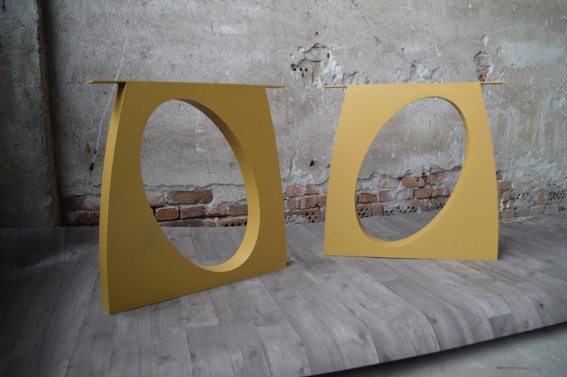 May include: Two gold metal table legs with an oval cutout in the center. The legs are standing on a wooden floor.