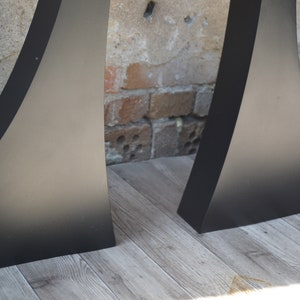 May include: Two black metal table legs with a curved design. The legs are standing on a light wood floor.