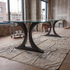 May include: A large rectangular table featuring a clear glass top supported by two dark gray metal bases. Each base has a distinct curved, crescent-like shape that flares outward at the bottom to provide stability for the expansive glass surface.