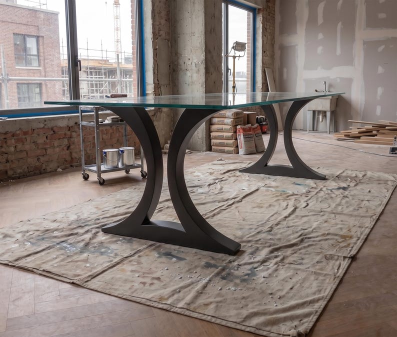 May include: A large rectangular table featuring a clear glass top supported by two dark gray metal bases. Each base has a distinct curved, crescent-like shape that flares outward at the bottom to provide stability for the expansive glass surface.