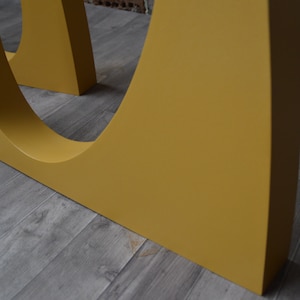 May include: Close-up of a yellow table leg with a curved design. The leg is made of wood and has a smooth finish.