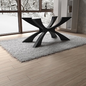 May include: A modern, black table base with a unique, geometric design. The base is made of metal and has multiple curved legs that intersect at the center. It sits on a gray, shaggy rug, with a snowy outdoor scene visible through a large window.