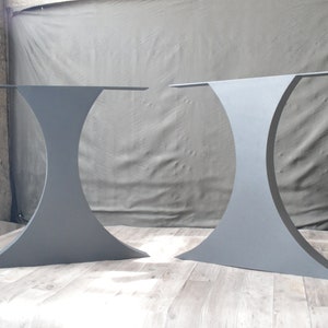 May include: Two gray metal table legs with a curved, hourglass shape. The legs are made of steel and have a matte finish.