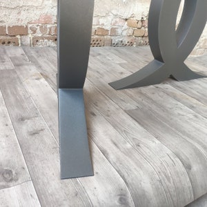 Puede incluir: Dos patas de mesa de metal negro con un dise&ntilde;o curvo &uacute;nico. Las patas se muestran sobre un suelo de imitaci&oacute;n de madera.