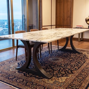 May include: A rectangular dining table featuring a polished white marble top with prominent grey veining. The table is supported by two curved, matte black metal pedestal bases that flare outward at the floor and the tabletop connection points.