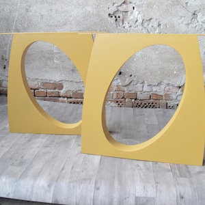May include: Two yellow metal table legs with oval cutouts. The legs are standing on a wood floor.