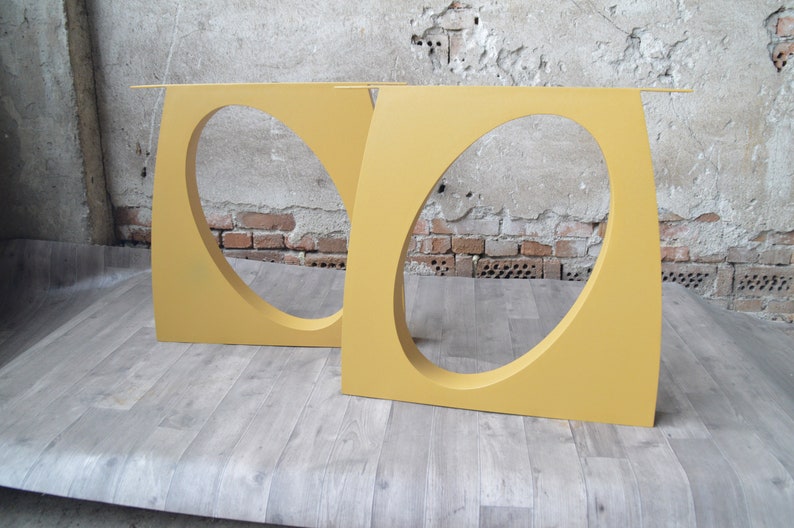 May include: Two yellow metal table legs with oval cutouts. The legs are standing on a wood floor.
