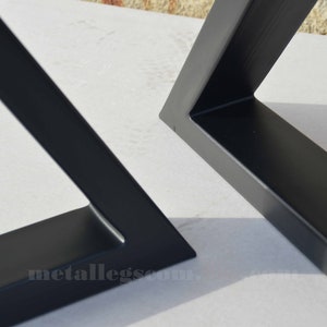 May include: Two black metal table legs, one in a triangular shape and the other in a square shape. The legs are made of thick metal and have a smooth, matte finish. The legs are shown on a white surface.