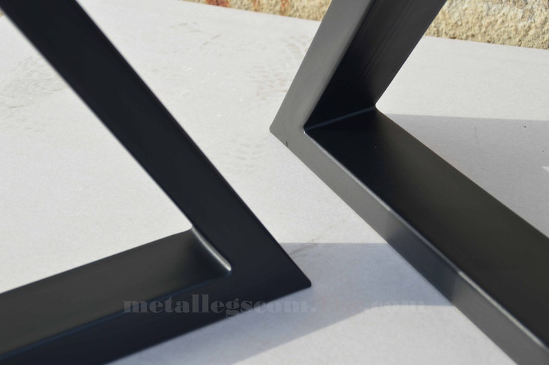 May include: Two black metal table legs, one in a triangular shape and the other in a square shape. The legs are made of thick metal and have a smooth, matte finish. The legs are shown on a white surface.