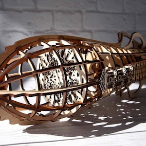May include: A wooden model of a steampunk airship with intricate details and a cage-like structure. The model is made of light brown wood and has a detailed design.