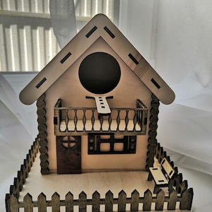 May include: A wooden birdhouse with a brown roof, a small balcony, and a picket fence. The birdhouse has a round entrance hole and a small window.
