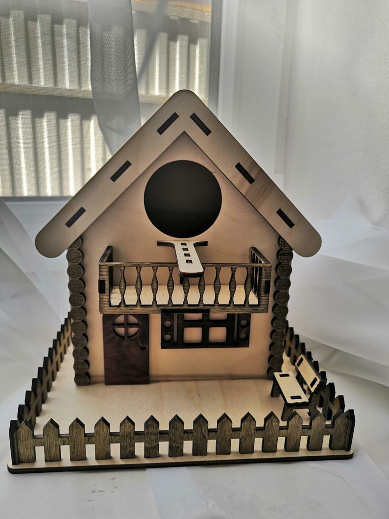 May include: A wooden birdhouse with a brown roof, a small balcony, and a picket fence. The birdhouse has a round entrance hole and a small window.