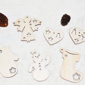 Puede incluir: Seis adornos navideños de madera en varias formas, incluyendo un ángel, una manopla, una media, un muñeco de nieve y dos corazones. Los adornos son todos blancos y tienen diseños recortados de estrellas, copos de nieve y árboles de Navidad.