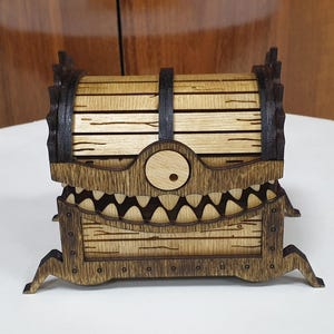 May include: A wooden treasure chest with a toothy monster face and a single eye. The chest is made of light and dark wood and has a hinged lid.