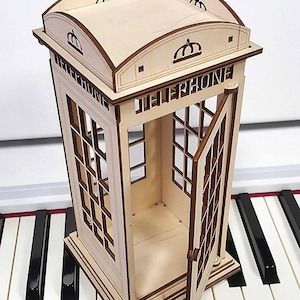 以下が含まれることがあります： 「TELEPHONE」の文字が刻まれた、木製の英国風電話ボックスのミニチュア。 ドアが開いており、内部が見えます。 モデルは薄茶色で、濃い茶色のトリムがあり、ピアノの上に置かれています。