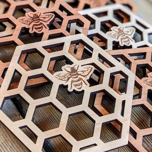 May include: Wooden coasters with a honeycomb design and a laser-cut bee. The coasters are made of light and dark brown wood. The hexagonal coasters are approximately 4 inches wide.