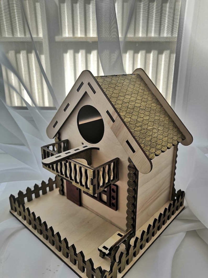 May include: A wooden birdhouse with a brown roof, a small balcony, and a picket fence. The birdhouse has a round entrance hole and a small window.