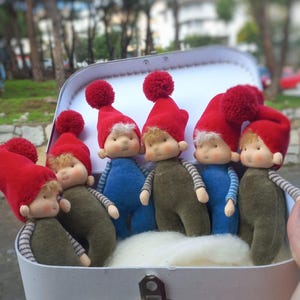 Puede incluir: Un grupo de muñecas hechas a mano en una maleta blanca. Las muñecas llevan sombreros rojos con pompones, monos azules o verdes y camisas a rayas. Las muñecas tienen la piel clara y el pelo rubio. La maleta está abierta.