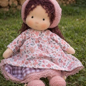 May include: A handmade cloth doll with a pink floral dress, matching bonnet, and crocheted shoes. The doll has brown hair and is sitting on green grass. The dress has a lace trim and a checkered pattern peeking out from underneath.
