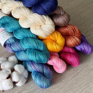 May include: A collection of multi-colored yarn skeins arranged in a fan shape, featuring shades of deep blue, cream, teal, yellow, brown, rust, purple, magenta, and pink, accompanied by several white cotton bolls with visible labels.