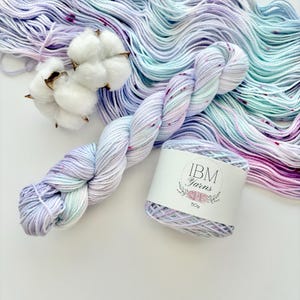 May include: A skein of hand-dyed yarn in shades of lavender, teal, and pink, with a speckled effect. A label reads "IBM Yarns" and "50g". Fluffy cotton bolls are also present. The yarn is ideal for knitting or crochet projects.