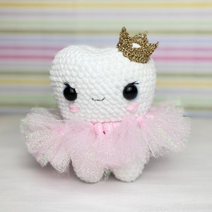 Op de afbeelding: Een witte gehaakte tand met een roze tule tutu en een gouden glitterkroon. De tand heeft een glimlachend gezicht met zwarte ogen.