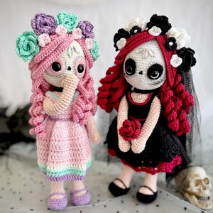 Könnte beinhalten: Zwei gehäkelte Puppen in rosa und schwarzen Kleidern mit Blumenkränzen. Die Puppen haben Schädelgesichter mit bemalten Details und halten Rosen.