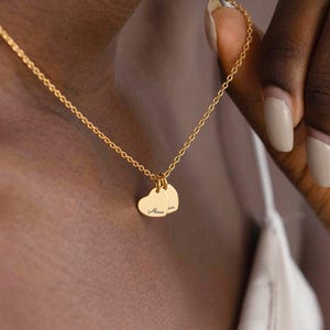 May include: A gold necklace featuring two heart-shaped pendants with engraved text. The pendants are suspended from a fine chain. The necklace is held by a person.