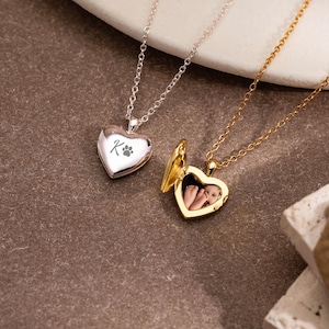 Puede incluir: Un collar con dije de corazón plateado grabado con la letra K y una huella de pata, junto a un collar con dije de corazón dorado abierto que muestra una pequeña fotografía de los pies de un niño, ambos con cadenas metálicas finas.