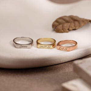 May include: Three metal rings with flat rectangular faces, each featuring custom engraving. The silver ring is engraved with the name Emma, the gold ring with the date 04.07.26, and the rose gold ring with the initials D & S in a cursive script font.