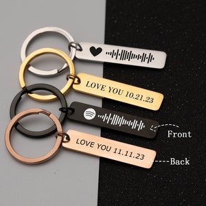 Pode incluir: Quatro chaveiros prateados com acabamento preto, dourado e ouro rosa. Cada chaveiro tem uma etiqueta retangular com um design único e o texto "LOVE YOU" seguido de uma data. A frente da etiqueta apresenta um logotipo do Spotify e um código de barras, enquanto a parte de trás apresenta um coração e um código de barras.