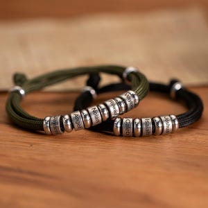 May include: Two personalized bracelets with silver beads and braided cord. One bracelet is olive green, the other is black. Each bracelet has multiple silver beads with names engraved on them. The bracelets are on a wooden surface.