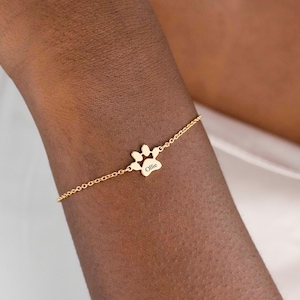 May include: A fine gold bracelet with a paw print pendant. The charm is inscribed with the name "Ollie". The bracelet is displayed on a person's wrist.