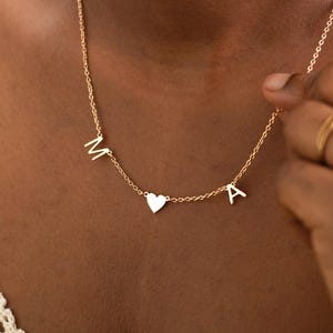 May include: A gold necklace featuring the letters "M" and "A" flanking a heart-shaped pendant. The necklace is worn around the neck. The person is wearing a white top.