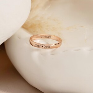 Puede incluir: Un anillo de banda de metal en tono oro rosa con un acabado liso y pulido. La superficie exterior está grabada con el texto 01/14/26 y los nombres Clara y Derek separados por un pequeño símbolo de corazón, creando una pieza de joyería personalizada.