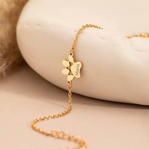 May include: A gold-colored bracelet featuring a paw print charm with the name "Maria" engraved on it. The bracelet is displayed on a white, curved surface, with a soft, neutral background. The chain is delicate and the overall aesthetic is elegant.