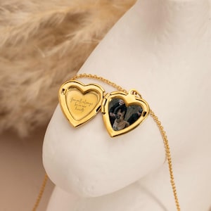 Puede incluir: Un collar con un colgante en forma de corazón dorado, abierto para revelar una fotografía. El medallón está grabado con las palabras "You will always be in our hearts". El collar se muestra sobre una superficie blanca.