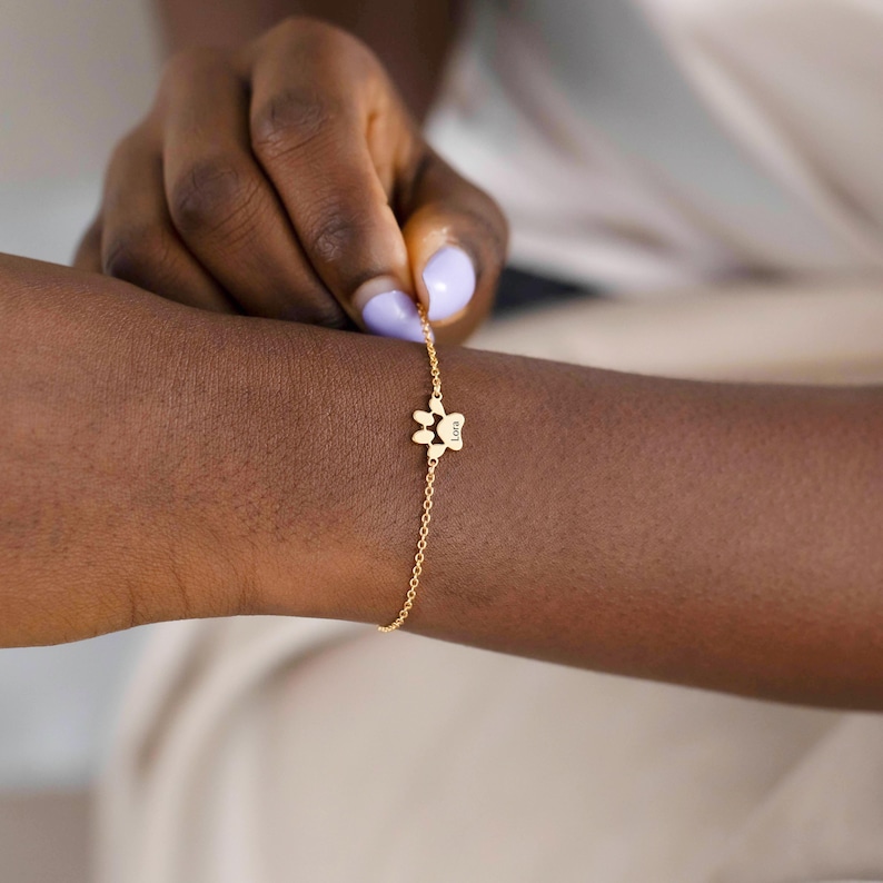 May include: A delicate gold-colored bracelet featuring a paw print charm. The charm has a small inscription. The bracelet is being held by a person with painted nails.