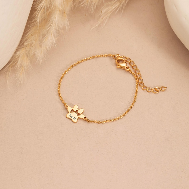May include: A gold-colored bracelet with a delicate chain and a paw print charm. The charm has the name "Freda" engraved on it. The bracelet is on a light pink surface.