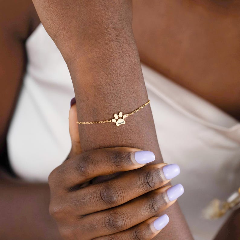 May include: A gold-colored bracelet featuring a paw print charm with the name "Annie" engraved on it. The delicate chain bracelet is worn on a wrist with light purple nail polish.
