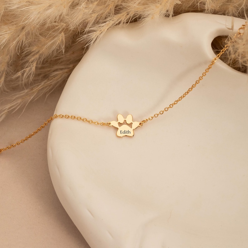 May include: A gold-colored bracelet featuring a paw print charm with the name "Edith" engraved on it. The charm is connected to a delicate chain. The bracelet is displayed on a white surface with dried plants in the background.
